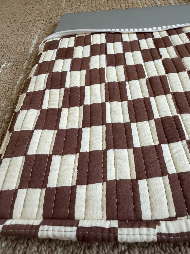May include: Close-up of a quilted fabric with a brown and cream checkerboard pattern. The fabric is stitched with a linear pattern. The edge of the fabric is visible, with a grey border.