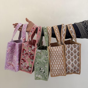 Könnte beinhalten: Eine Auswahl an kleinen Tragetaschen mit verschiedenen floralen und geometrischen Mustern. Die Taschen sind in Rosa-, Grün- und Brauntönen gehalten und haben passende gemusterte Griffe und Verzierungen. Die Taschen werden von einer Hand gehalten.