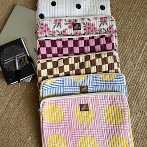 May include: A selection of quilted laptop sleeves with diverse patterns. Designs include white with black polka dots, pink floral, burgundy checkerboard, brown and cream checkerboard, blue striped, and pink with lemon slices. Each sleeve has a zip closure.