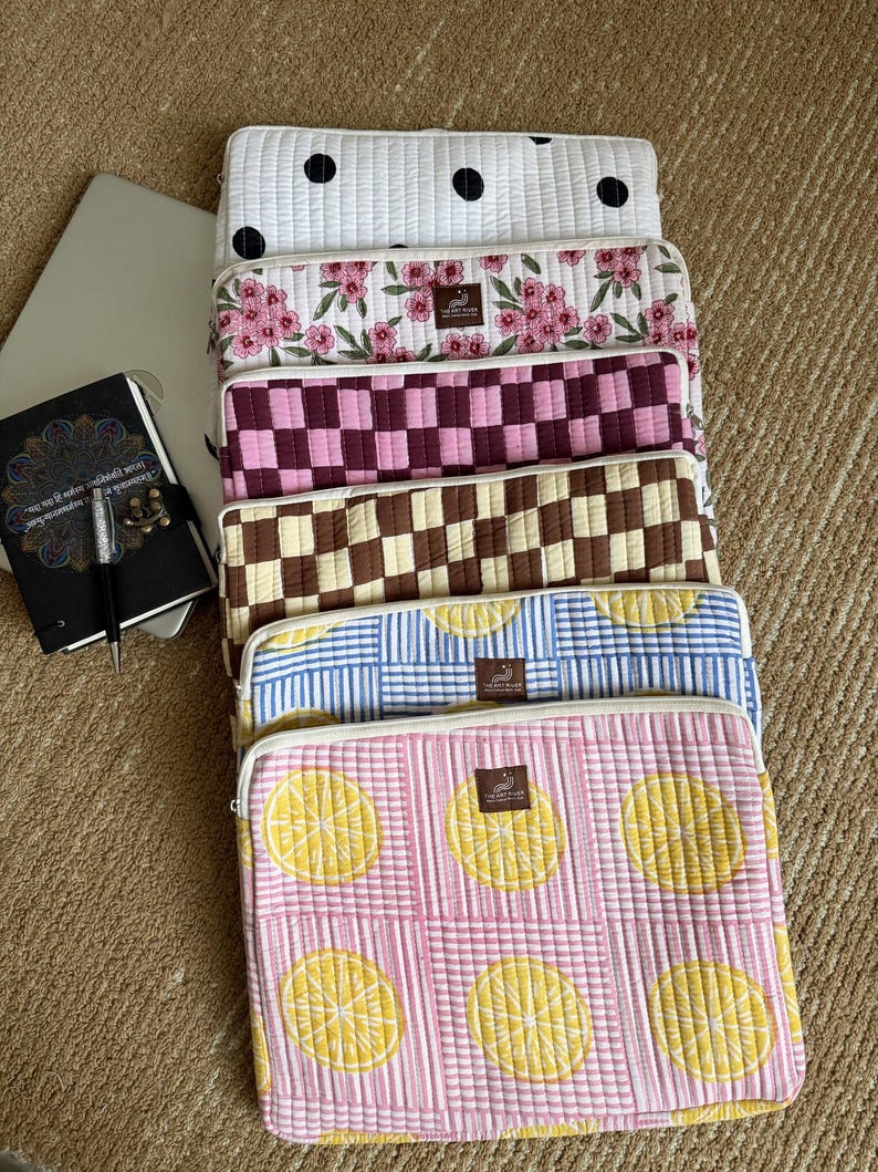 May include: A selection of quilted laptop sleeves with diverse patterns. Designs include white with black polka dots, pink floral, burgundy checkerboard, brown and cream checkerboard, blue striped, and pink with lemon slices. Each sleeve has a zip closure.