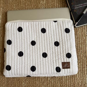 May include: White quilted laptop sleeve with a black polka dot pattern. The sleeve has a zip closure and a small brown label with text. A laptop is partially visible inside the sleeve. A notebook and pen are in the background.