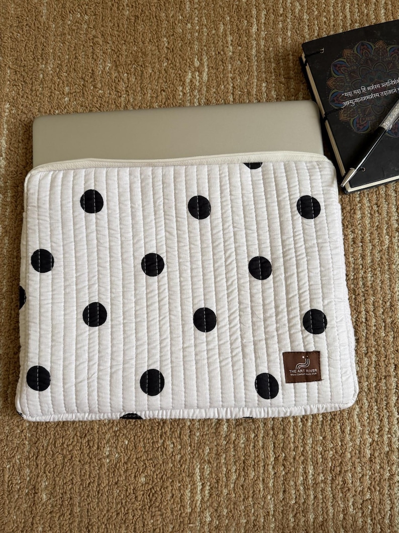 May include: White quilted laptop sleeve with a black polka dot pattern. The sleeve has a zip closure and a small brown label with text. A laptop is partially visible inside the sleeve. A notebook and pen are in the background.