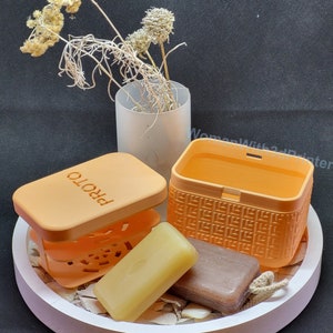 Könnte beinhalten: Zwei orangefarbene Seifenschalen mit weißem Hintergrund. Eine Schale hat die Aufschrift "PROTO" auf der Oberseite. Die andere Schale hat ein gewebtes Muster. Zwei Seifenstücke befinden sich in den Schalen.