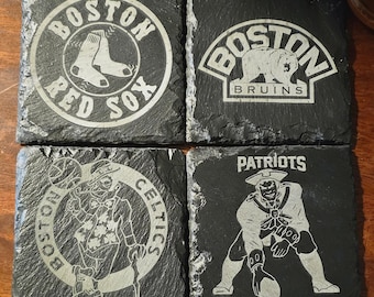 May include: Four square slate coasters featuring Boston sports team logos: Red Sox, Bruins, Celtics, and Patriots. Each coaster has a unique design etched in a light color against the dark slate.