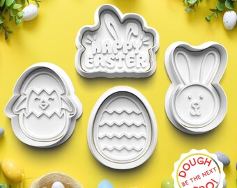 May include: Four white plastic cookie cutters on a yellow background. The cutters are shaped like a bunny, an egg with a wavy pattern, a chick in an egg, and a banner that says "Happy Easter".
