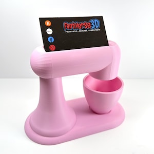 May include: A pink 3D-printed business card holder in the form of a stand mixer. A black business card is in the holder, with the text "Fantiverse3D" and social media icons. The holder includes a base, a bowl, and a mixer arm.