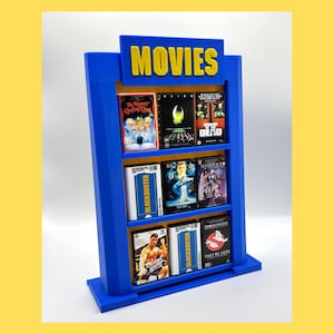 May include: A blue miniature movie shelf with the word "MOVIES" in yellow. The shelf holds several movie cases, including "The Muppet Christmas Carol," "Alien," and "Dawn of the Dead."