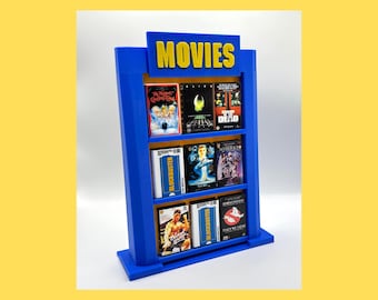May include: A blue miniature movie shelf with the word "MOVIES" in yellow. The shelf holds several movie cases, including "The Muppet Christmas Carol," "Alien," and "Dawn of the Dead."