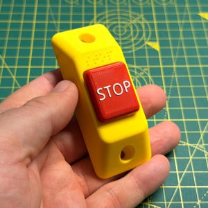May include: A bright yellow rectangular housing featuring a prominent red square button centred on the front. The button has the word STOP embossed in white letters. There are circular mounting holes at the top and bottom of the yellow base.