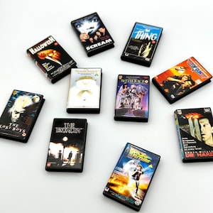 Puede incluir: Una colecci&oacute;n de cintas VHS en miniatura con t&iacute;tulos de pel&iacute;culas cl&aacute;sicas como "Halloween", "Scream", "The Thing" y "Regreso al Futuro". Las cintas son negras con ilustraciones y texto coloridos. Otros t&iacute;tulos incluyen "Beetlejuice" y "Die Hard".