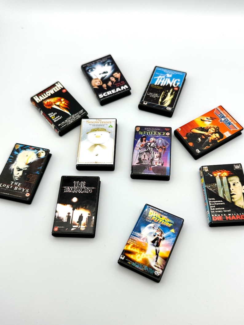 Puede incluir: Una colecci&oacute;n de cintas VHS en miniatura con t&iacute;tulos de pel&iacute;culas cl&aacute;sicas como "Halloween", "Scream", "The Thing" y "Regreso al Futuro". Las cintas son negras con ilustraciones y texto coloridos. Otros t&iacute;tulos incluyen "Beetlejuice" y "Die Hard".