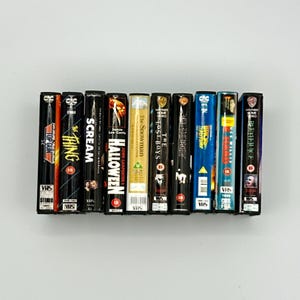 Puede incluir: Una colecci&oacute;n de cintas VHS vintage con t&iacute;tulos de pel&iacute;culas como "Top Gun", "The Thing", "Scream" y "Halloween". Las cintas est&aacute;n dispuestas en fila, mostrando sus coloridas lomos y las ic&oacute;nicas ilustraciones de las pel&iacute;culas. Las cintas miden aproximadamente 18 cm de alto.