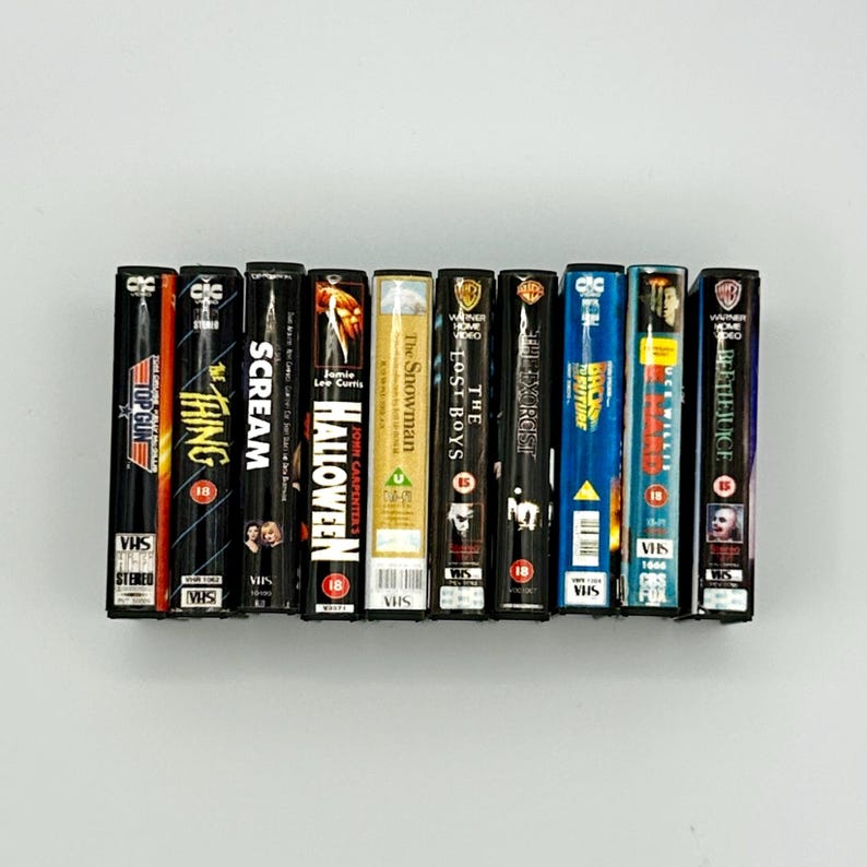 Puede incluir: Una colecci&oacute;n de cintas VHS vintage con t&iacute;tulos de pel&iacute;culas como "Top Gun", "The Thing", "Scream" y "Halloween". Las cintas est&aacute;n dispuestas en fila, mostrando sus coloridas lomos y las ic&oacute;nicas ilustraciones de las pel&iacute;culas. Las cintas miden aproximadamente 18 cm de alto.