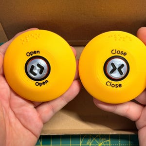 May include: Two circular yellow buttons with black text and symbols. The left button features the word Open and a bracket symbol, while the right button features the word Close and a bracket symbol. Both buttons include raised braille dots.