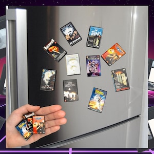 Puede incluir: Imanes de pel&iacute;culas VHS en miniatura, con t&iacute;tulos como "Halloween" y "Regreso al futuro", se muestran en un refrigerador. Una mano sostiene algunos m&aacute;s. El fondo presenta un dise&ntilde;o retro de los a&ntilde;os 80.