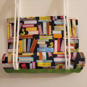 May include: A fabric swing seat with a black background covered in colorful book illustrations. The books have titles like "Dictionary", "History", and "Chemistry". White rope is attached for hanging, and the bottom edge is trimmed with green fabric.