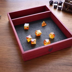 May include: A rectangular red wooden dice tray featuring a black textured leatherette base and a separate storage compartment. Six translucent orange and yellow polyhedral dice are scattered inside the tray, with a small engraved icon on the front.