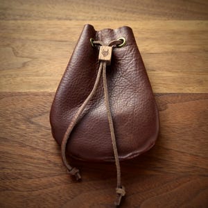 May include: A dark brown leather drawstring pouch featuring a textured surface, brass-toned eyelets, and a small square wooden bead engraved with a bull head logo that secures the thin leather cord closure.