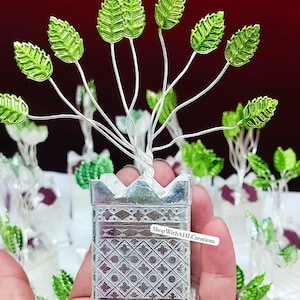 May include: A silver metal decorative tree with green leaves. The tree is in a square pot with a crown-like top and a detailed pattern. The text "ShopWithAHLCreations" is visible.