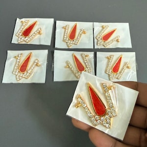 May include: Several gold-colored trident-shaped ornaments with red centers and clear crystal accents are displayed. Each ornament is packaged on a small, square, white card. One is held by a hand.