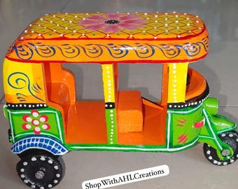 May include: A colorful, hand-painted wooden auto-rickshaw toy. The vehicle features a yellow roof with a floral design, orange and green body, and black wheels. The text "ShopWithAHLCreations" is visible.
