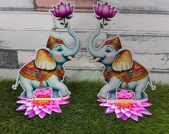 May include: Two decorative elephant figurines with light blue bodies, ornate gold and jewel-toned accents, and a pink lotus flower held in their trunks. The elephants stand on pink lotus flower bases, set against a backdrop of faux grass and a wood-paneled wall.