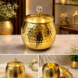 May include: A round, gold-colored metal canister with a hammered texture and a removable lid. The lid features a clear, faceted crystal-like knob at the top. The canister has a wide, bulbous body and a polished, reflective metallic finish.