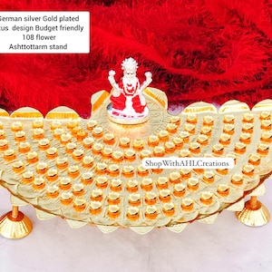 May include: A gold-plated lotus design Ashtottarm stand with a small figurine. The stand has a semi-circular shape with numerous small, flower-shaped accents. The background is a vibrant red fabric. Text on the image reads "German silver Gold plated lotus design Budget friendly 108 flower Ashtottarm stand".