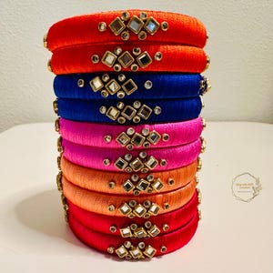 May include: A stack of colorful silk thread bangles with decorative mirror embellishments. The bangles are in shades of red, blue, pink, and orange.