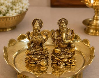 Puede incluir: Dos estatuas de metal en tono dorado de las deidades Lakshmi y Ganesha, sentadas sobre bases de loto. Las figuras están colocadas juntas sobre una bandeja redonda de latón con bordes festoneados y un acabado pulido brillante.