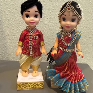 May include: Two dolls dressed in traditional Indian attire. The doll on the left wears a red and gold shirt and white pants with gold trim. The doll on the right wears a red and blue sari with ornate jewelry and a headpiece. Both dolls are on decorative stands.