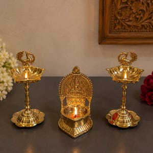 May include: Three gold-colored brass oil lamps with lit flames. Two lamps feature tall pedestals with peacock finials, while the central lamp has a low, ornate backplate with embossed figures. Each lamp has a polished, reflective metallic finish.