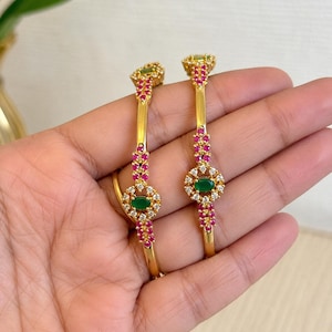 May include: Two gold bangles featuring a central oval green stone surrounded by small white stones and accented with rows of small pink stones. The metal bands are smooth and polished, with the decorative elements centered on the outer surface.