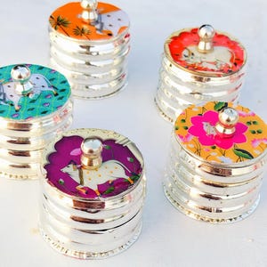 May include: Five small, silver-colored, round containers with lids. Each lid features a different colorful design with a central knob. The lids display various illustrations, including animals and floral patterns. The containers are stacked with a ribbed design.