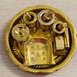 May include: A circular gold-colored metal tray containing several brass-toned items, including three cylindrical containers with lids, a rectangular block, a small rod, and a shovel-shaped piece with engraved figures, partially wrapped in clear plastic.