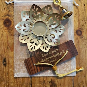May include: A decorative brass flower-shaped candle holder, a small brass bell, and a wooden tag with text are displayed on a sheer bag. The candle holder has a central well for a candle. The wooden tag has text in a script font.