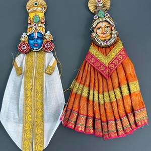May include: Two decorative figures featuring detailed faces and ornate headpieces. The left figure has blue skin, wearing a white garment with gold trim. The right figure has a light complexion, wearing a pleated orange and pink dress with gold accents.