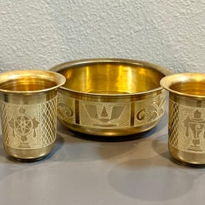 May include: A set of three brass items consisting of one round bowl and two matching tumblers. Each piece features intricate etched detailing, including geometric cross-hatching and symbolic motifs on the exterior surfaces of the metal vessels.