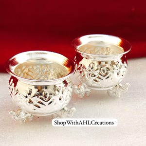 May include: Two silver-colored decorative bowls with ornate floral patterns and small elephant-shaped feet. The bowls have a wide, slightly flared rim and are displayed on a white surface against a red backdrop. The text "ShopWithAHLCreations" is visible.