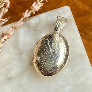 Op de afbeelding: Een ovaalvormige zilveren medaillonhanger met een gedetailleerd gegraveerd bladpatroon op de voorzijde. Het medaillon is voorzien van een gepolijst metalen oogje aan de bovenzijde voor bevestiging aan een ketting en heeft een glanzende afwerking.