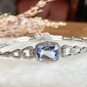 May include: A silver bracelet with an Art Deco design. The bracelet features a large, rectangular, light blue gemstone at its center. The chain links are intricately detailed with small, decorative patterns.