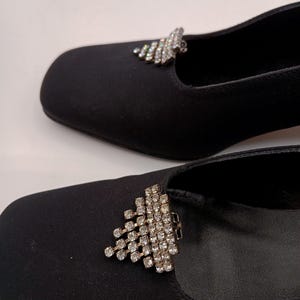May include: A pair of black satin shoes featuring a square toe design. Each shoe is adorned with a triangular-shaped decorative clip encrusted with clear, sparkling rhinestones positioned on the top front of the vamp.