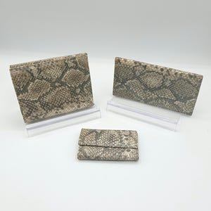 May include: Three faux snakeskin wallets in varying sizes. The wallets are displayed on clear acrylic stands against a white background. The pattern is a mix of tan, brown, and gray, resembling a snake's skin. The wallets appear to be made of a textured material.