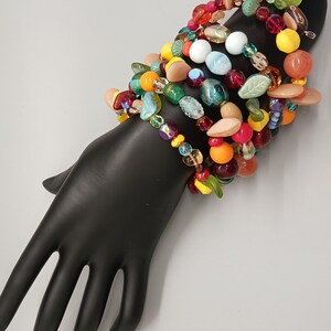 May include: A stack of colorful bracelets with glass beads, leaves, and other decorative elements. The bracelets are arranged on a black mannequin hand.