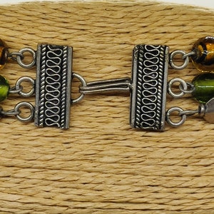 May include: A silver bracelet with three strands of beads. The beads are green, brown, and yellow. The bracelet has a silver clasp with a decorative design.