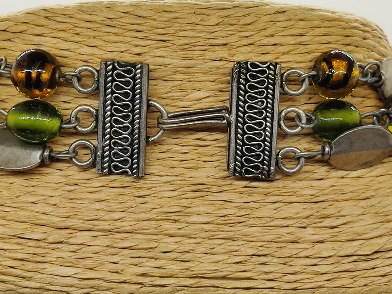 May include: A silver bracelet with three strands of beads. The beads are green, brown, and yellow. The bracelet has a silver clasp with a decorative design.