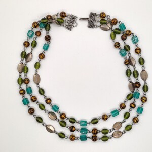 May include: A three-strand necklace with green, brown, and silver beads. The necklace has a silver clasp.