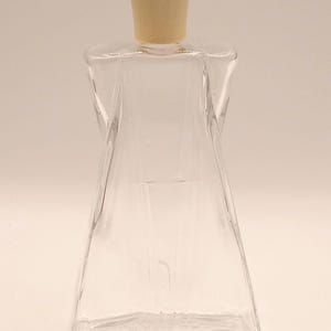May include: A clear glass perfume bottle with a cream-colored stopper. The bottle has a unique, twisted design and a flared base. The stopper is cylindrical. The bottle is empty and ready to be filled.