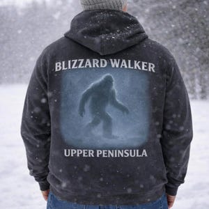 May include: Dark gray hoodie with the words "BLIZZARD WALKER" above a silhouette of a Bigfoot-like creature. Below the image is the text "UPPER PENINSULA". The hoodie is worn in a snowy outdoor setting.
