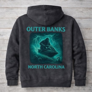 May include: Dark grey hooded sweatshirt with the text "OUTER BANKS NORTH CAROLINA" in teal. The back displays a teal graphic of a Sasquatch riding a jet ski in stormy waters.